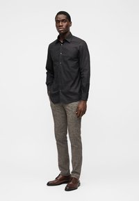 Camisa preta com botões, em tecido texturizado, combinada com calças de espinha de peixe castanhas e sapatos de couro escuro, posando contra um fundo simples.