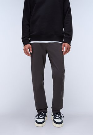 Mann trägt schwarzen Sweatshirt, dunkelgraue Hosen und weiße und schwarze Sneaker und steht vor einem schlichten hellen Hintergrund.
