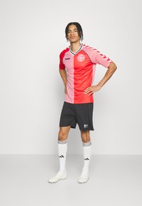 Miesjalkapalloilija seisoo käsivarret lanteilla, päällään punavalkoraidallinen pelipaita, mustat shortsit, valkoiset polvenkorkuiset sukat ja valkoiset nappikset.