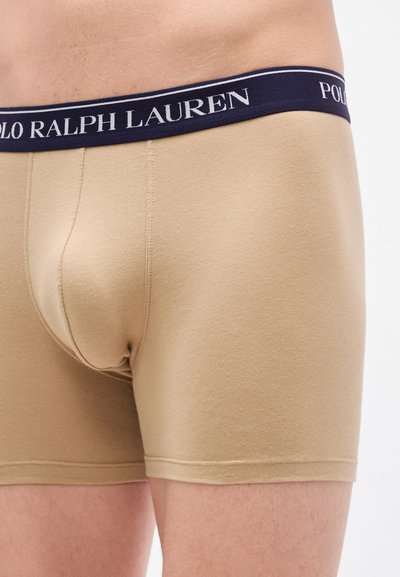 Μπεζ εφαρμοστά μποξέρ με μπλε ναυτικό ζωνάρι μέσης που φέρει το λογότυπο "POLO RALPH LAUREN". Μαλακό ύφασμα με λεία υφή και επίπεδες ραφές.
