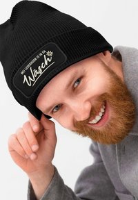 Schwarze Strickmütze mit einem rechteckigen Patch, auf dem weißer Text und ein Blumenmuster zu sehen sind. Der Text lautet "MEI LEDERHOSN IS IN DA Wäsche."