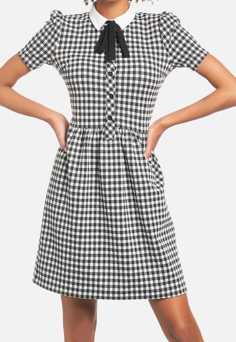 Musta-valkoinen ruudullinen mekko, jossa on lyhyet puffihihat, valkoinen kaulus, napit edessä ja laskostettu hame.