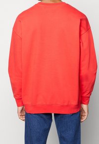 Rotes Sweatshirt mit Rundhalsausschnitt, überschnittenen Schultern und gerippten Bündchen. Glatte Textur und lockere Passform, kombiniert mit blauen Jeans.