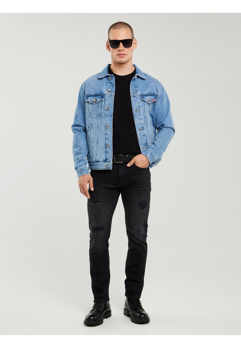 Homme portant des lunettes de soleil noires, une veste en denim bleue, un t-shirt noir, un jean noir déchiré et des bottes noires, se tenant devant un fond uni.