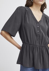 Graues, kurzärmeliges Blusenshirt mit V-Ausschnitt, das über eine Knopfleiste und einen elastischen Bund für einen gerafften Look verfügt. Glattes Material mit leichtem Glanz.