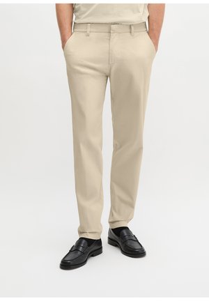 Mann trägt beige, gerade geschnittene Hose mit Seitentaschen und schwarze Leder-Loafers vor weißem Hintergrund.