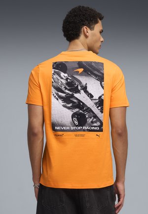 Uomo che indossa una maglietta arancione con grafica di un'auto da corsa in bianco e nero e il testo "Never Stop Racing" sulla schiena.