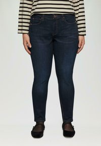 Dunkelblaue Skinny-Jeans mit glatter Textur, Fünf-Taschen-Design und orangefarbenen Nähten. Kombiniert mit einem schwarzen flachen Schuh.