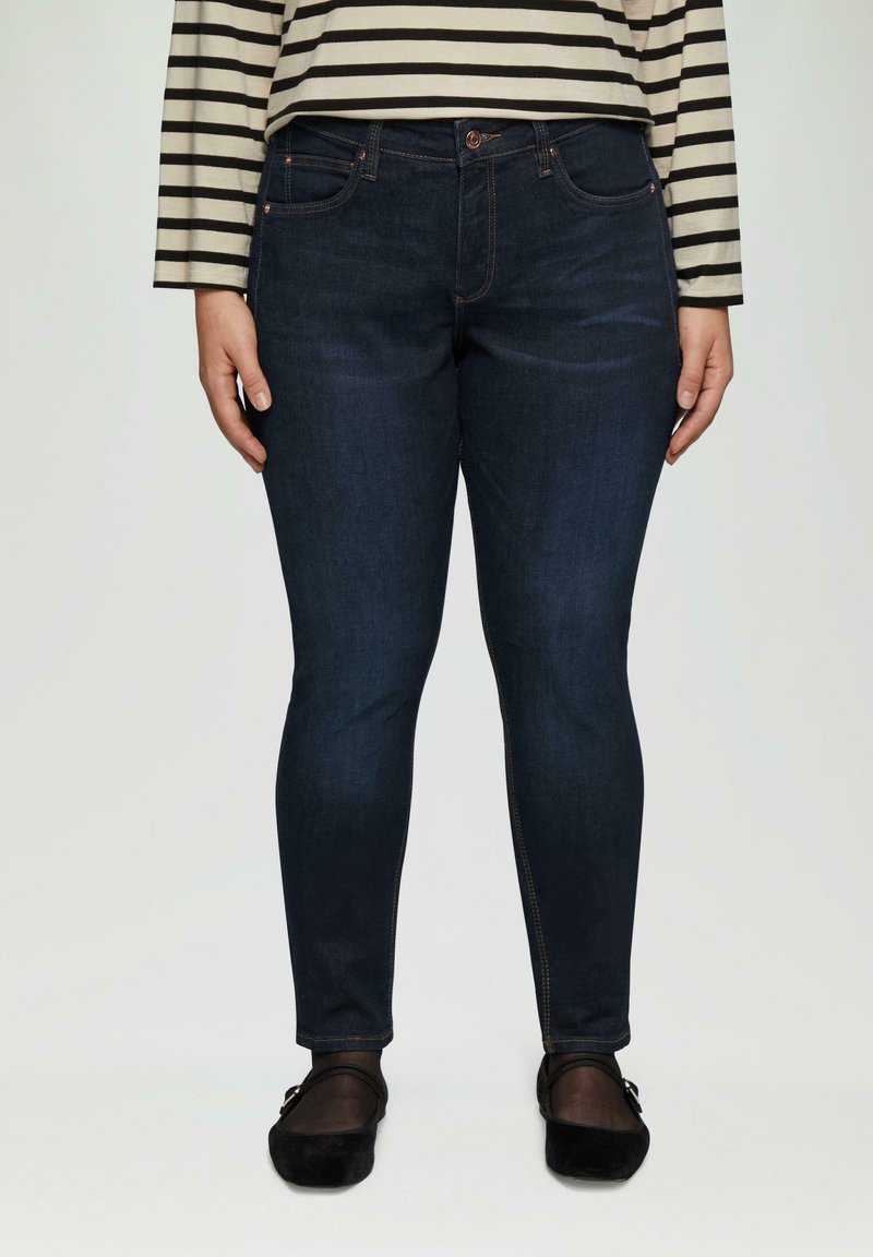 Dunkelblaue Skinny-Jeans mit glatter Textur, Fünf-Taschen-Design und orangefarbenen Nähten. Kombiniert mit einem schwarzen flachen Schuh.