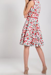 Weißes Blumenkleid mit roten Rosen, ärmelloses Design, taillierter Mieder, ausgestellter Rock und einem passenden roten Gürtel. Getragen mit nuden High Heels.