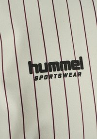Tkanina odzieży sportowej Hummel z kremowym tłem i pionowymi bordowymi paskami. Logo jest wyraźnie widoczne na czarno w centrum.