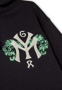 Sudadera negra con un destacado logotipo bordado en color crema y verde, rodeado de intrincados motivos de hojas que realzan las letras.