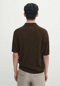 Brązowa dzianinowa koszulka polo z krótkim rękawem, z prążkowanym kołnierzykiem i mankietami, o luźnym kroju i gładkiej fakturze, noszona na jasnych spodniach.