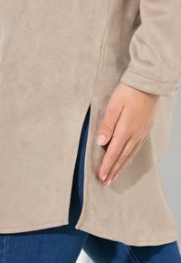 Beige Tunika aus samtigem Material mit langen Ärmeln, seitlichen Schlitzen und einer glatten Textur, kombiniert mit blauen Denim-Jeans. Nahaufnahme einer Hand auf der Tunika.