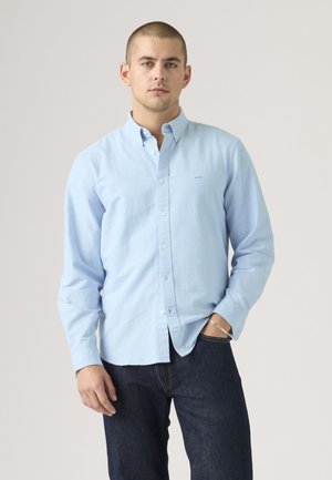 Uomo che indossa una camicia celeste con bottoni e jeans scuri, in piedi con una mano in tasca davanti a uno sfondo semplice.
