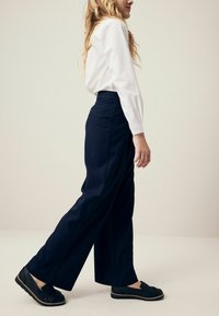 Tamsiai mėlynos, plačios kelnės iš lygios medžiagos, derinamos su baltu marškinėliu. Komplektą papildo paprasti juodi batai su tekstūruotu dizainu.