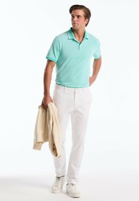 Polo shirt vert menthe avec une texture, pantalon blanc, et une veste beige clair drapée sur un bras, complétée par des baskets blanches.