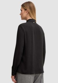 Camicia nera a maniche lunghe con collo alto, texture liscia e sottile accento metallico sul retro. Vestibilità ampia e design semplice.