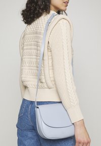 Světlomodrá kožená kabelka přes rameno s hladkou texturou, zakřivenou klopou a nastavitelným páskem. N nošena přes pletený smetanový svetr.