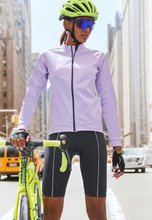 Light purple cycling jacket with black shorts; green helmet and gloves; cyclist holds a yellow bike with black and blue accents in a city.