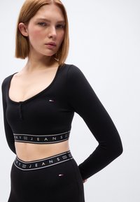 Μαύρο crop top με μακριά μανίκια, στρογγυλή λαιμόκοψη, κλείσιμο με κουμπιά και ελαστική ταινία στο κάτω μέρος με το κείμενο "TOMMY JEANS".