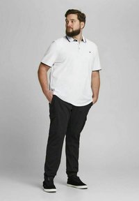 Biała koszulka polo z czarnym obramowaniem, zestawiona z czarnymi spodniami cargo. Model stoi swobodnie, ręce w kieszeniach, ma na sobie czarne sneakersy.