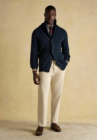 Marineblauer gerippter Cardigan mit Schalkragen und Knöpfen, über einem gemusterten Hemd getragen, kombiniert mit beigen Hosen und braunen Slippern.