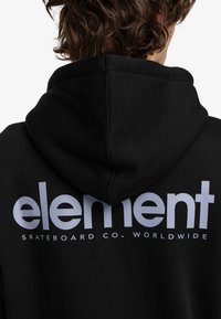 Sudadera negra con capucha que presenta un gran logotipo impreso con "element" y "SKATEBOARD CO. WORLDWIDE" en blanco. El material parece suave y texturizado.