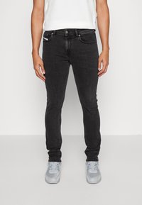 Jeans slim fit negros de tejido de mezclilla con cinco bolsillos, cierre de botón metálico y ligero desvanecimiento. Combinados con zapatillas deportivas gris claro.