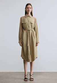 Φόρεμα πουκάμισο σε χακί από απαλό ύφασμα, με γιακά, δύο τσέπες στο στήθος, κλείσιμο με κουμπιά και ζώνη στη μέση δεμένη. Το τελείωμα είναι ελαφρώς καμπυλωτό.