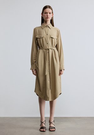Khakifarbenes Hemdblusenkleid aus glattem Stoff mit Kragen, zwei Brusttaschen, Knopfleiste und gebundenem Taillengürtel. Der Saum ist leicht geschwungen.