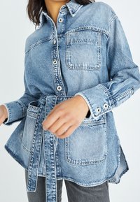 Ljusblå denimskjorta med skärp i midjan, långa ärmar, knäppning, bröstficka och detaljer med rå fåll. Har en avslappnad passform.