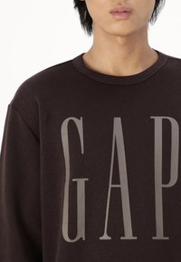 Hnědá bavlněná mikina s velkým šedým logem "GAP" na přední straně, kulatým výstřihem a volným střihem. Hladká textura, neformální design.
