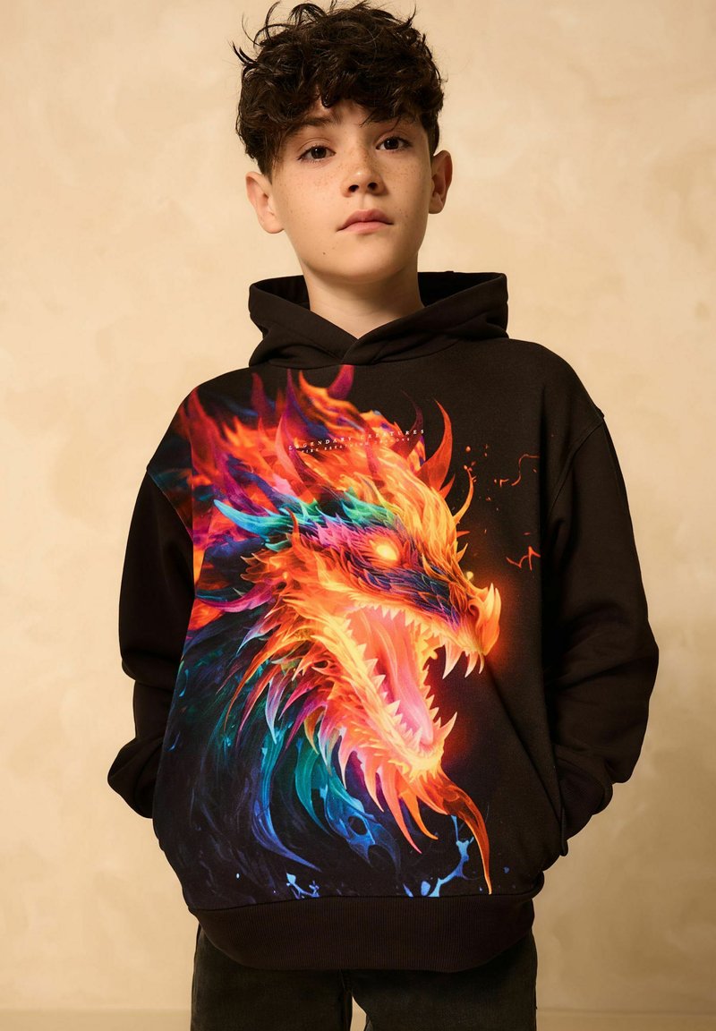 Junge mit dunklen lockigen Haaren trägt schwarzen Hoodie mit lebhaftem feurigem Drachenmotiv und steht vor einem schlichten beigen Hintergrund.