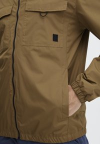 Brown lightweight jacket with a front zipper, two chest pockets, elastic cuffs, and a small logo patch on the left side.