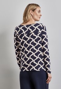 Granatowa bluzka z długim rękawem z geometrycznym kremowym wzorem, okrągłym dekoltem i luźnym krojem z tylnym przeszyciem. Miękka tekstura materiału.