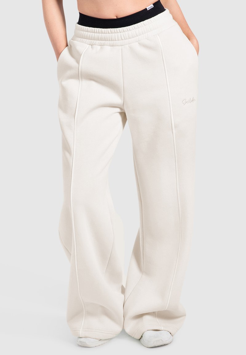 Pantalon de survêtement blanc ample avec coutures visibles et poches latérales, porté avec une ceinture noire visible et des baskets blanches en dessous.