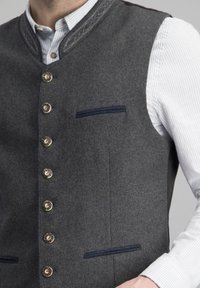 Graham Wolle Weste mit hohem Kragen, verziert mit dekorativen Nähten, Messingknöpfen und einem kleinen blauen Taschendetail auf der linken Seite.
