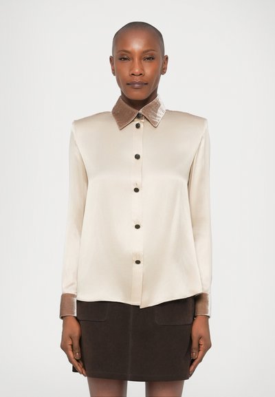 Chemise en satin beige avec un col en velours marron, manches longues et sept boutons noirs sur le devant. Texture douce, coupe ajustée.