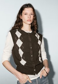 Brown cardigan with white argyle pattern, button front, ribbed hem, worn over a white long-sleeve shirt, paired with denim.