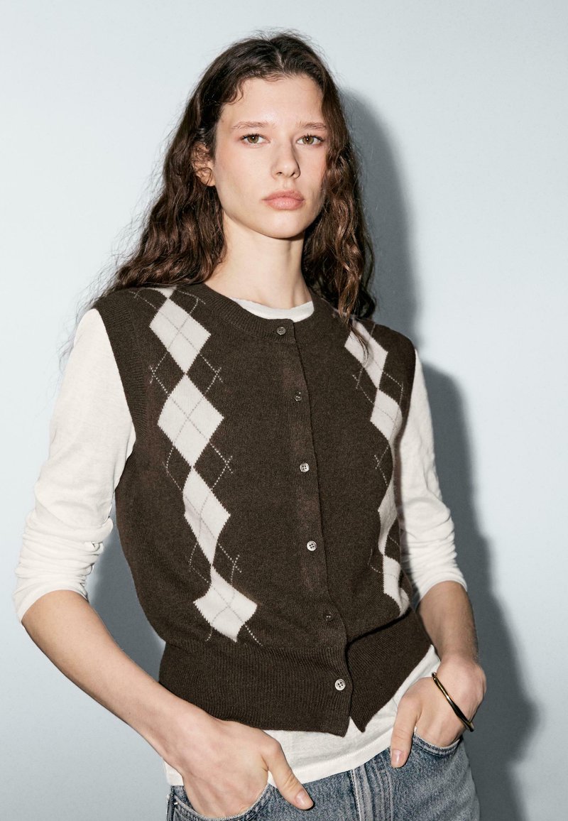 Brown cardigan with white argyle pattern, button front, ribbed hem, worn over a white long-sleeve shirt, paired with denim.