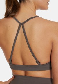 Harmaa urheiluliivi, jossa on ribattua tekstiiliä, urheilullinen takakappale ristiin menevillä hihnoilla ja säädettävät hakaset takana varmistamassa tukevan istuvuuden.