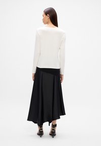 Langärmeliges weißes Oberteil, kombiniert mit einem schwarzen Satinrock mit asymmetrischem Saum, abgerundet durch schwarze hohe Schuhe.