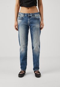 Mujer vistiendo jeans de pierna recta descoloridos en azul claro, top negro corto y zapatos Mary Jane negros, de pie contra un fondo claro y liso.