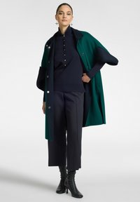 Groene en marineblauwe oversized jas met zilveren knopen over een marineblauw top en gemusterde donkere broeken, gecombineerd met zwarte stiletto laarzen.