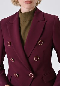 Blazer bordeaux doppiopetto con dettagli in metallo dorato, rever a punta e silhouette aderente, indossato sopra un dolcevita verde.