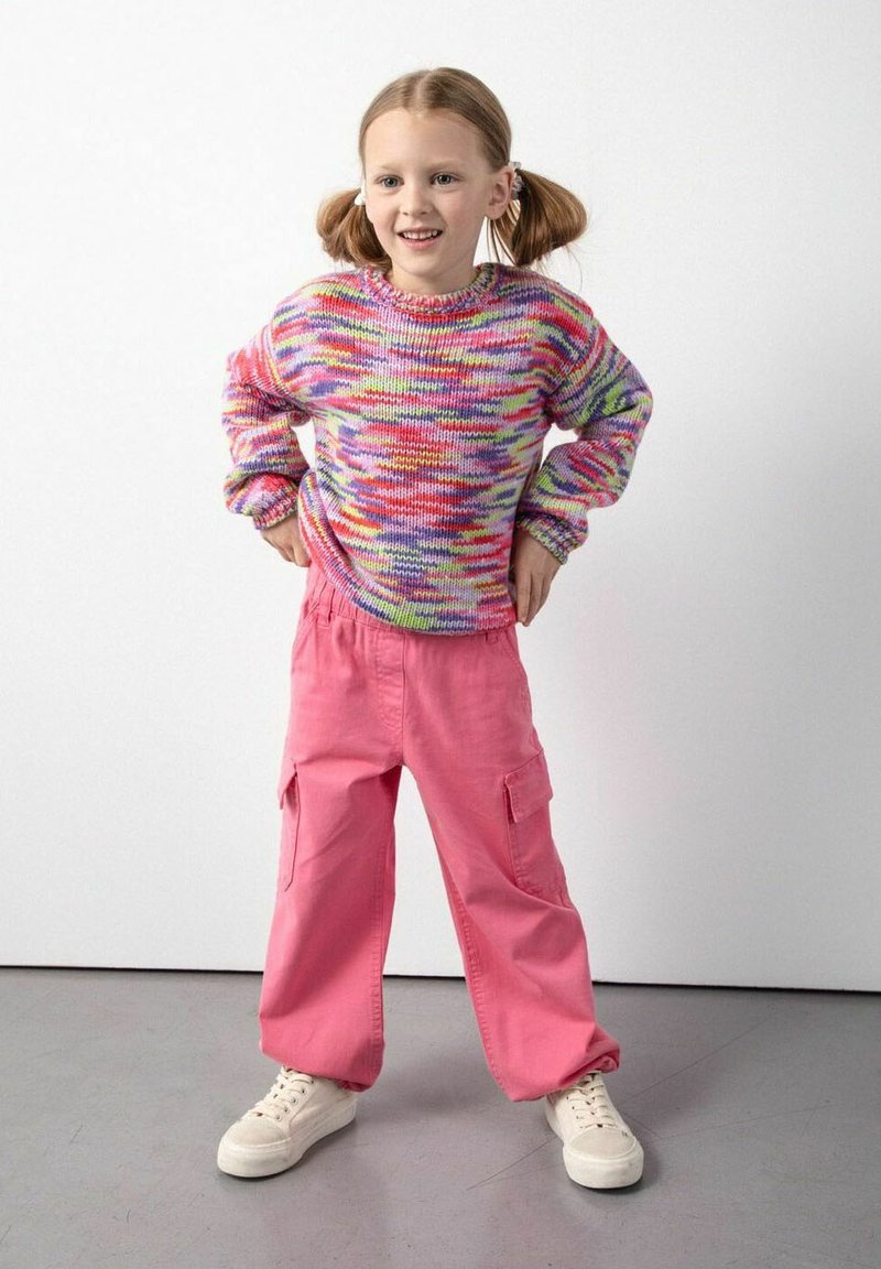 Młoda dziewczynka z warkoczami, nosząca kolorowy sweter z wełny, różowe spodnie cargo i białe trampki, stojąca z rękami na biodrach.
