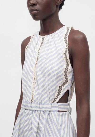 Femme portant une robe sans manches blanche à rayures bleu clair avec un galon en dentelle festonné et une ceinture découpée sur le côté.