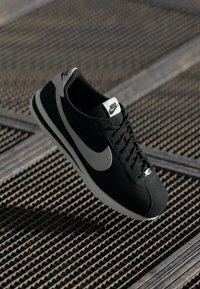 Sort sneakers med grå detaljer, lavet af ruskind og mesh-materiale, rund tå, flade snørebånd, og Nike-logo på tungen og siden.