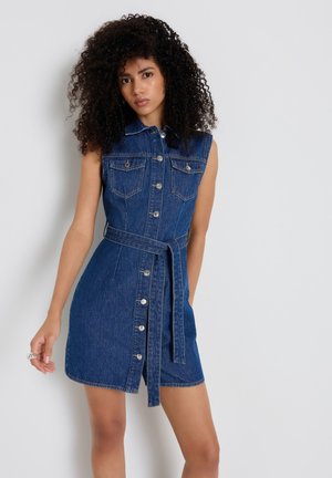 Frau mit lockigem Haar, die ein ärmelloses blaues Jeanskleid mit Knöpfen vorne, Taschen und einem Taillengürtel trägt, steht vor einer schlichten Wand.